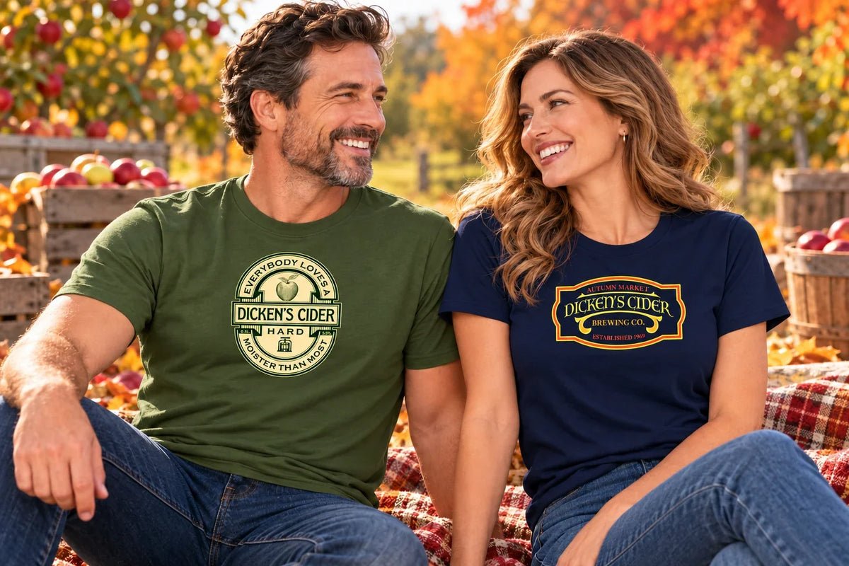 Two adults wearing Dickens Cider T-shirts with vintage cider-label graphics at an autumn apple orchard.