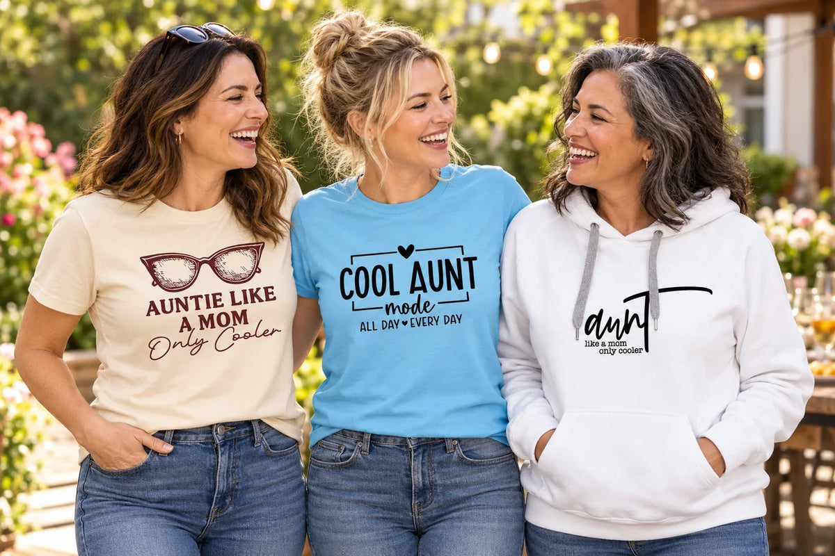 Three women wearing Aunt and Auntie graphic T-shirts and a hoodie at a garden gathering.